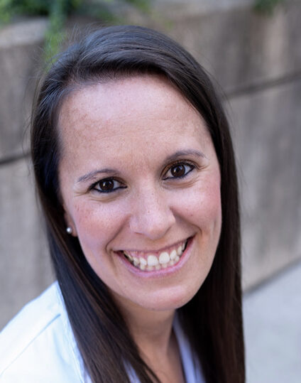 Dr. Waters smiles. She is outdoors, with a blurred concrete wall and greenery in the background.